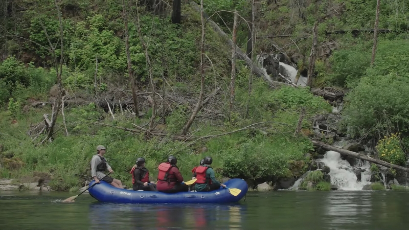 A group in helmets and life jackets rafts along a river in Oregon surrounded by lush greenery and a small waterfal