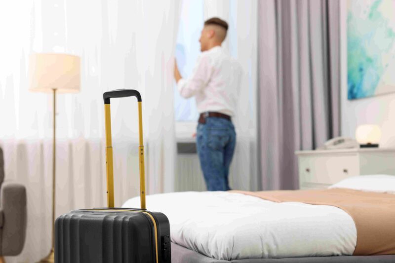 Suitcase beside a hotel bed while a traveler stands near the window inside a hotel room