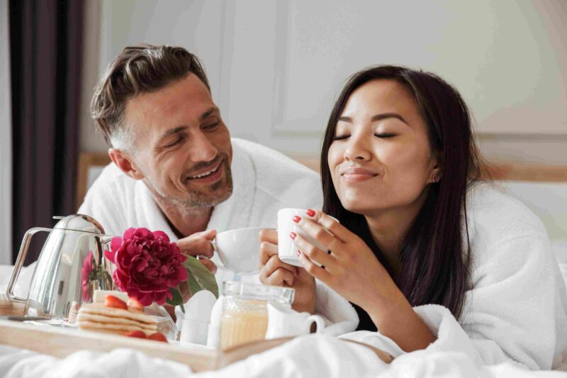 Couple in hotel robes enjoying breakfast in bed with coffee and flowers