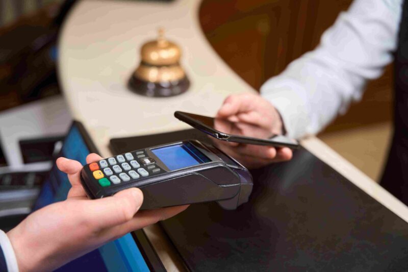Guest making a contactless payment at a hotel front desk with card terminal and phone