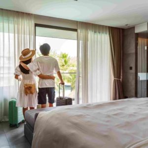 Couple standing in a hotel room with luggage while looking out toward a balcony view
